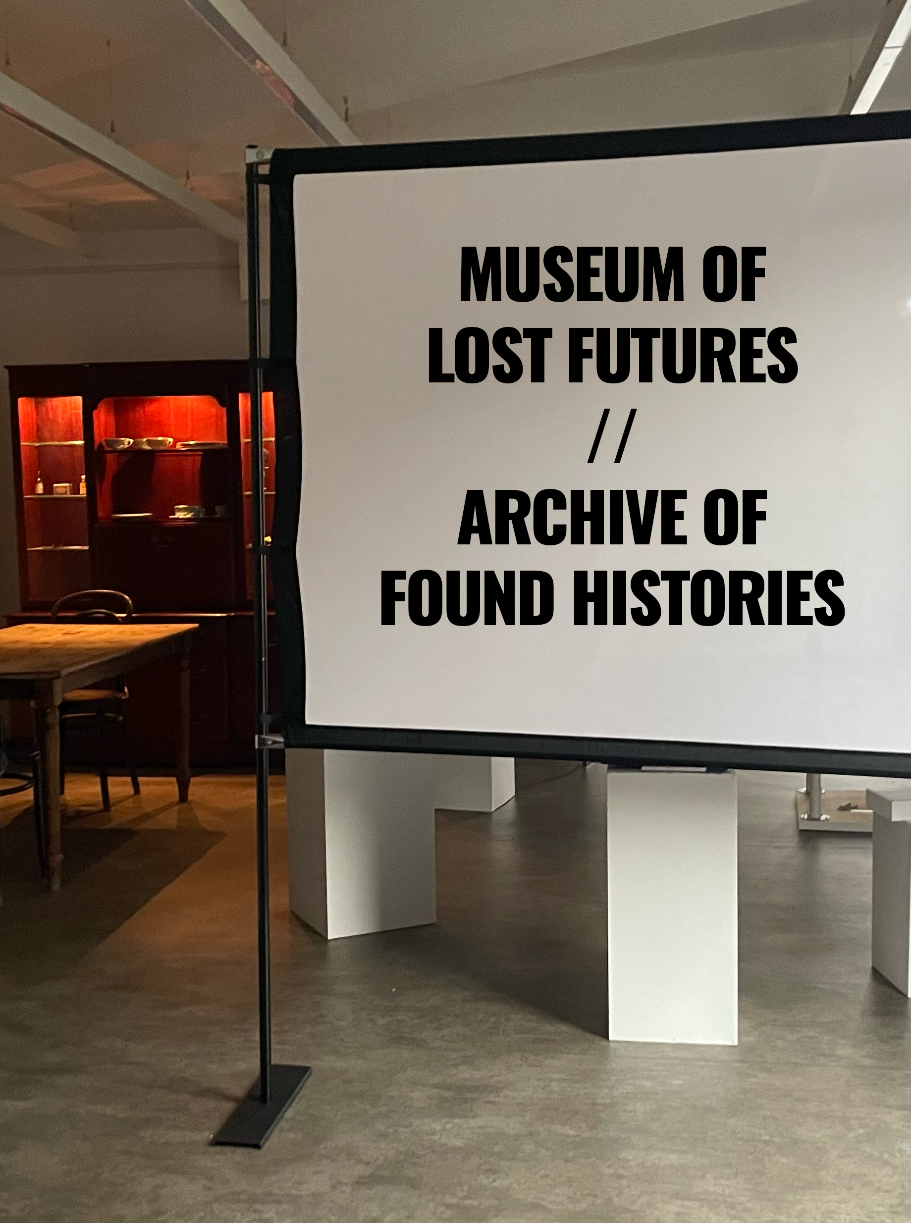 A gallery. Foreground: a screen with the words "Museum of Lost Futures". Background: a dining table and Welsh dressers.