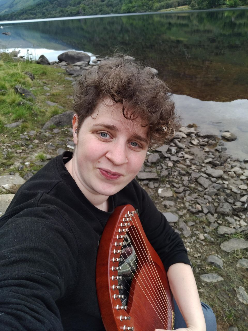 A short-haired person holding a musical instrument by a stream. They look mischievous.