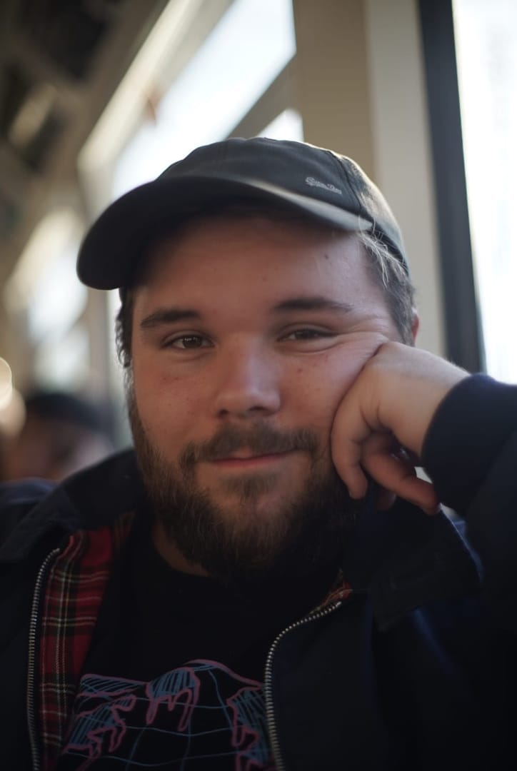 A softly-focused photo of a white non-binary person with a beard on a train. They're wearing a cap and look quite happy.