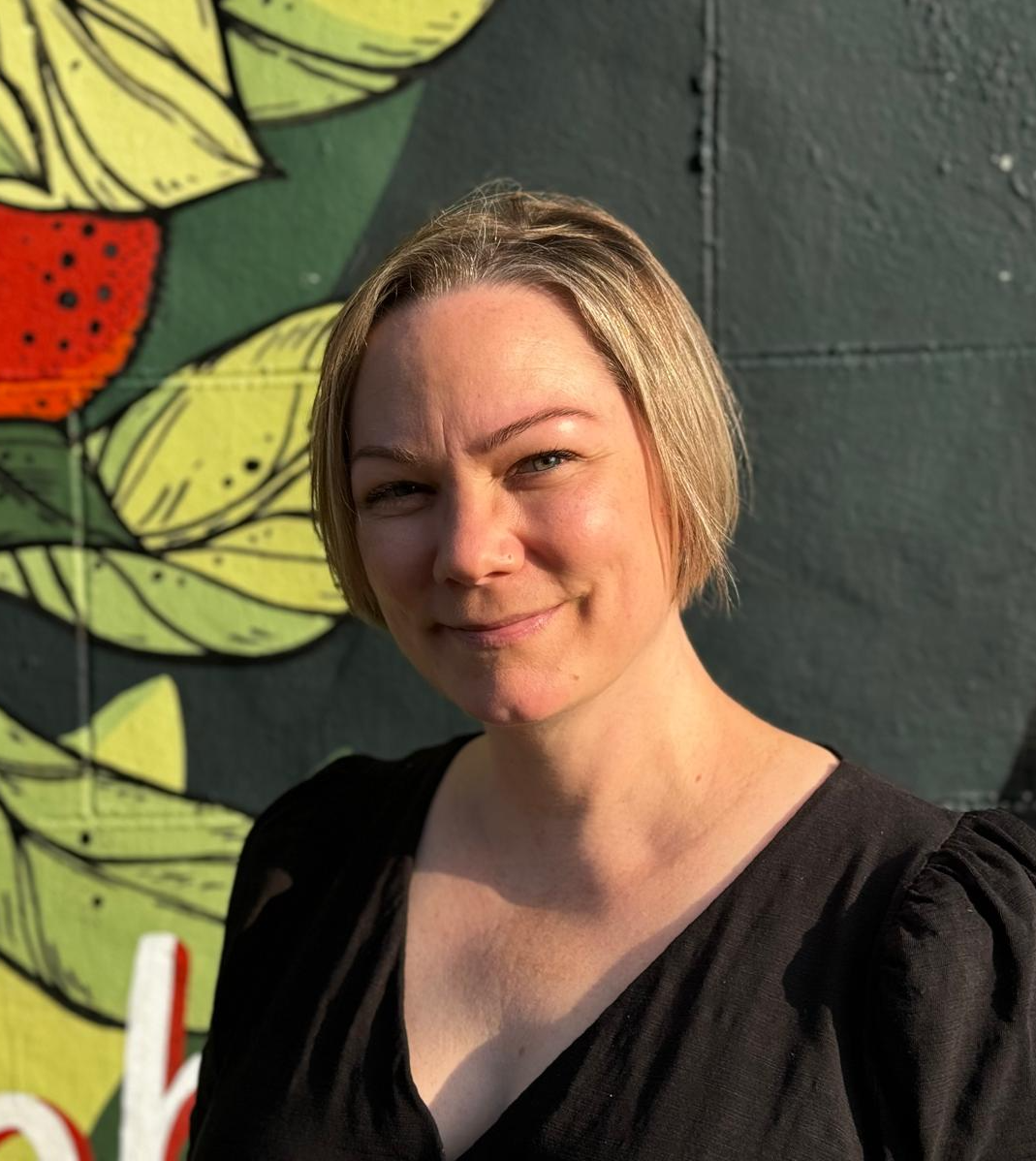 A white non-binary person standing against a wall painted with an image of a plant bearing fruit. They are smiling and staring into the sun, which is making them squint a little.