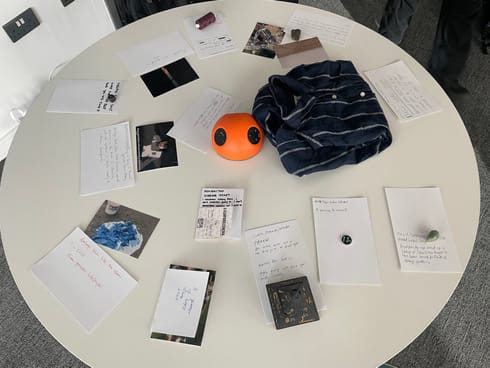 A round table with items on it. Each item has a handwritten museum placard. The items include clothing, esotera, photos.