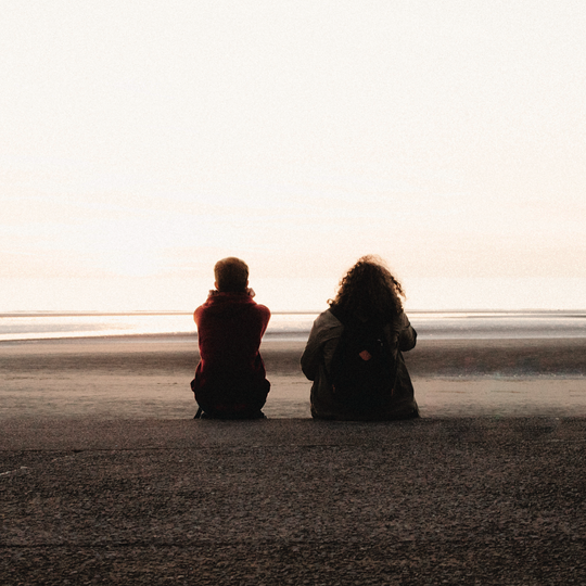 A picture of a young person and an adult, sitting on a beach staring into the distance together.