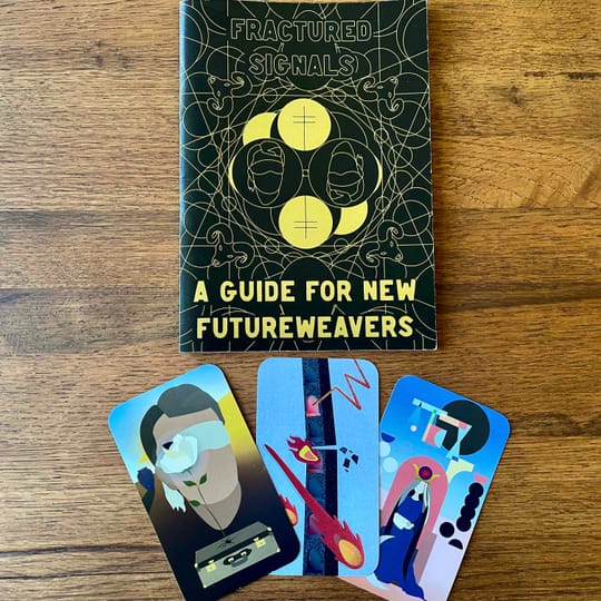 3 tarot cards on a wooden table underneath a black guidebook that says "fractured signals: a guide for new futureweavers"
