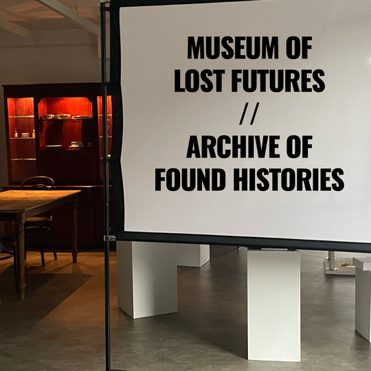 A gallery. Foreground: a screen with the words "Museum of Lost Futures". Background: a dining table and Welsh dressers.